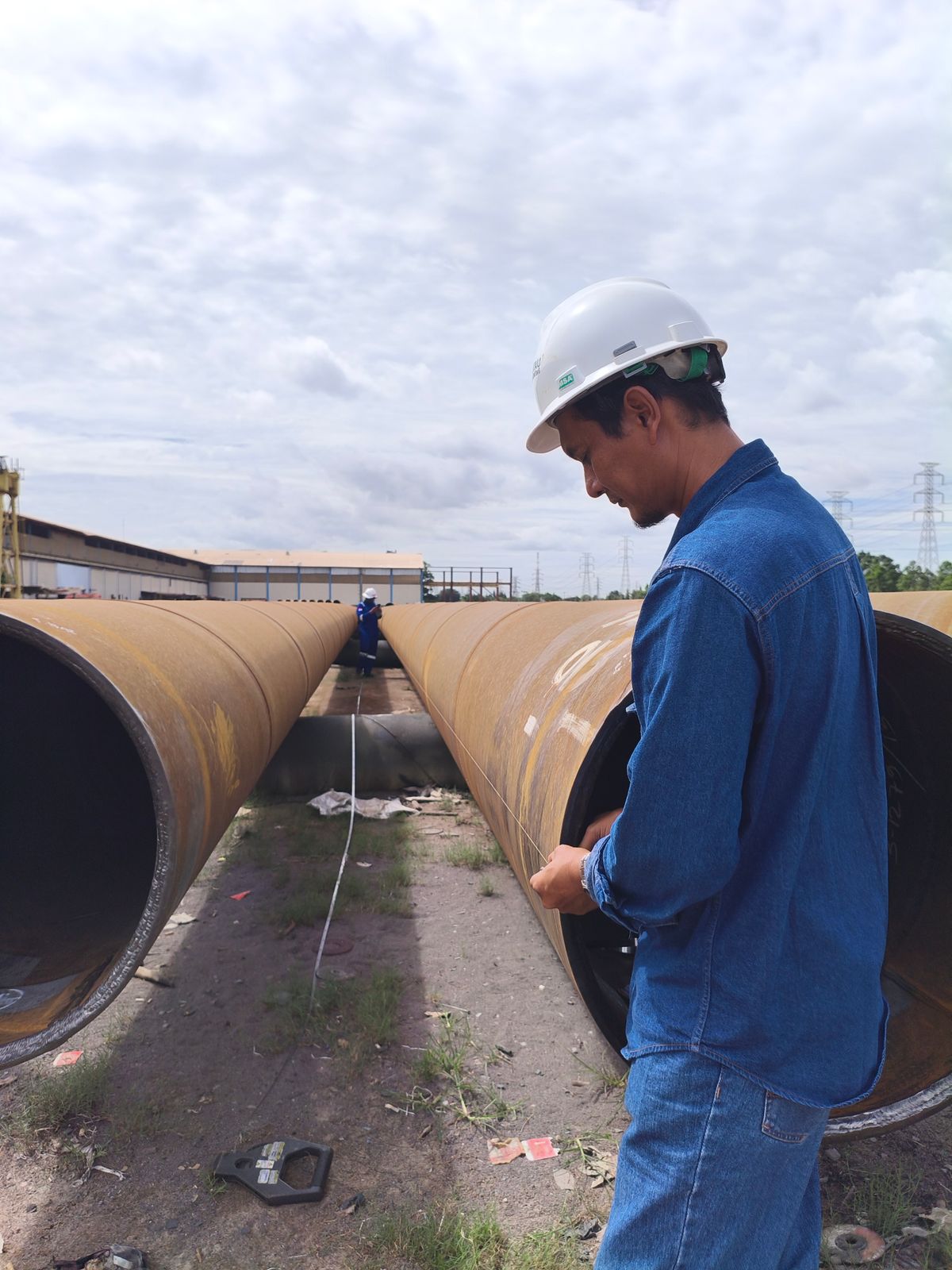 A man in a hard hat measuring a length of large steel pipe in a pipe yard